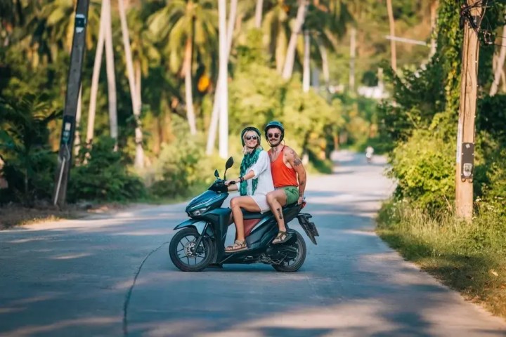 Two people on a scooter on a tropical road with palm trees.