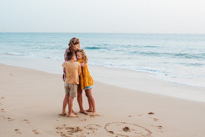 Woman and two children hugging on a sandy beach near ocean waves.