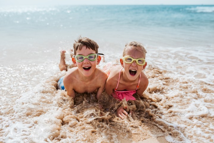 Two kids with goggles lying in shallow ocean waves, smiling and playing on the beach.