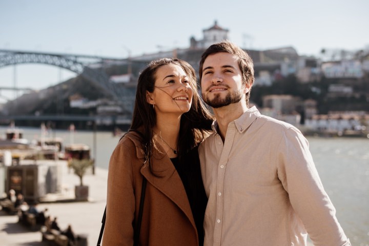 Smiling couple by a river, bridge and cityscape in the background on a sunny day.