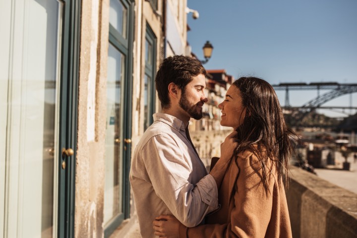 Couple smiling at each other near a building with blue doors, bridge in background.