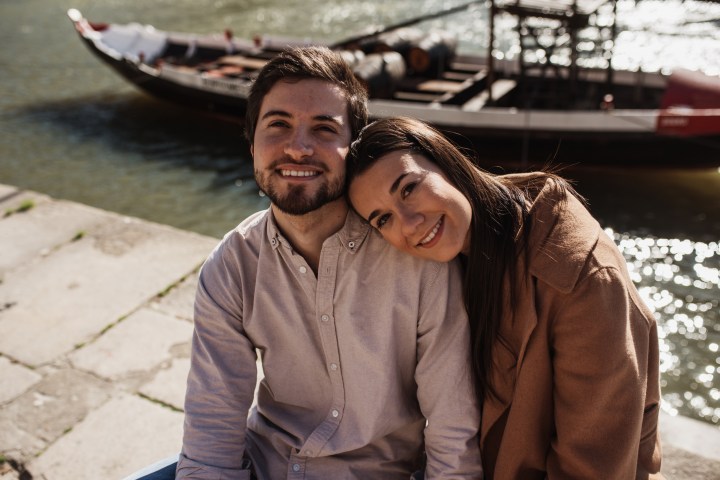 Smiling couple sitting by a river, with a boat in the background.