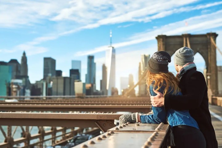 Couple in winter clothing embraces on a bridge, overlooking a city skyline under a blue sky.
