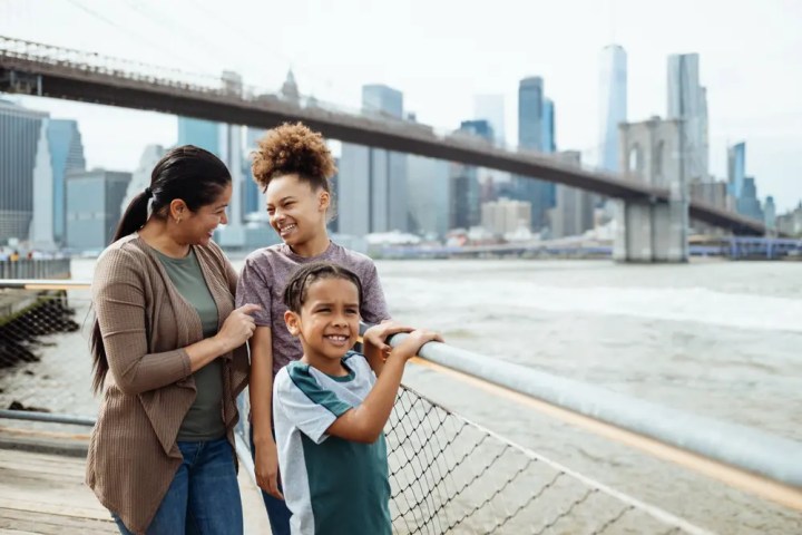 Smiling family of three near river with city skyline and bridge in background.